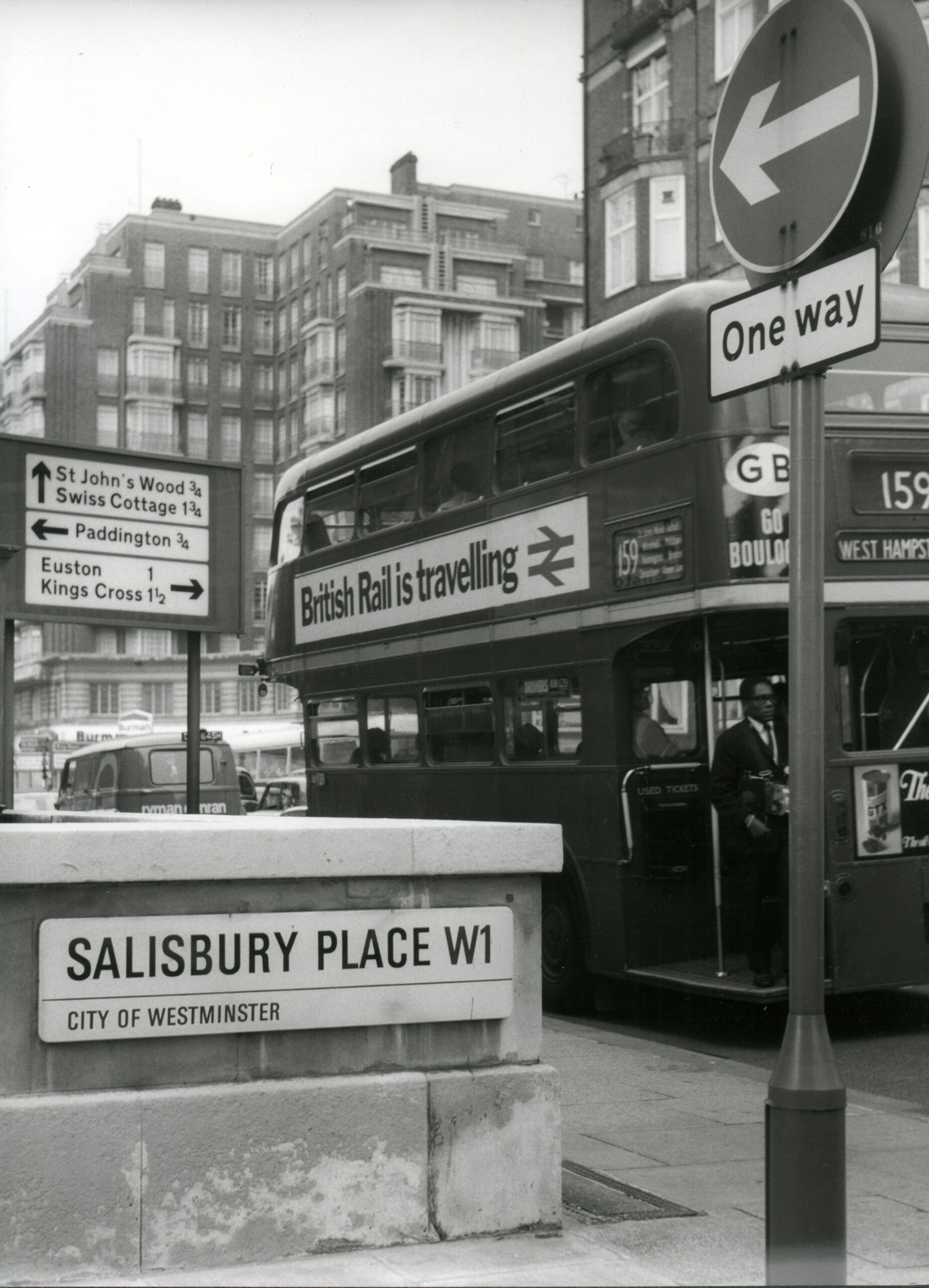 a black and white image of a London bus next to a subway entrance. There are lots of road signs and street signs which are present in the photo.