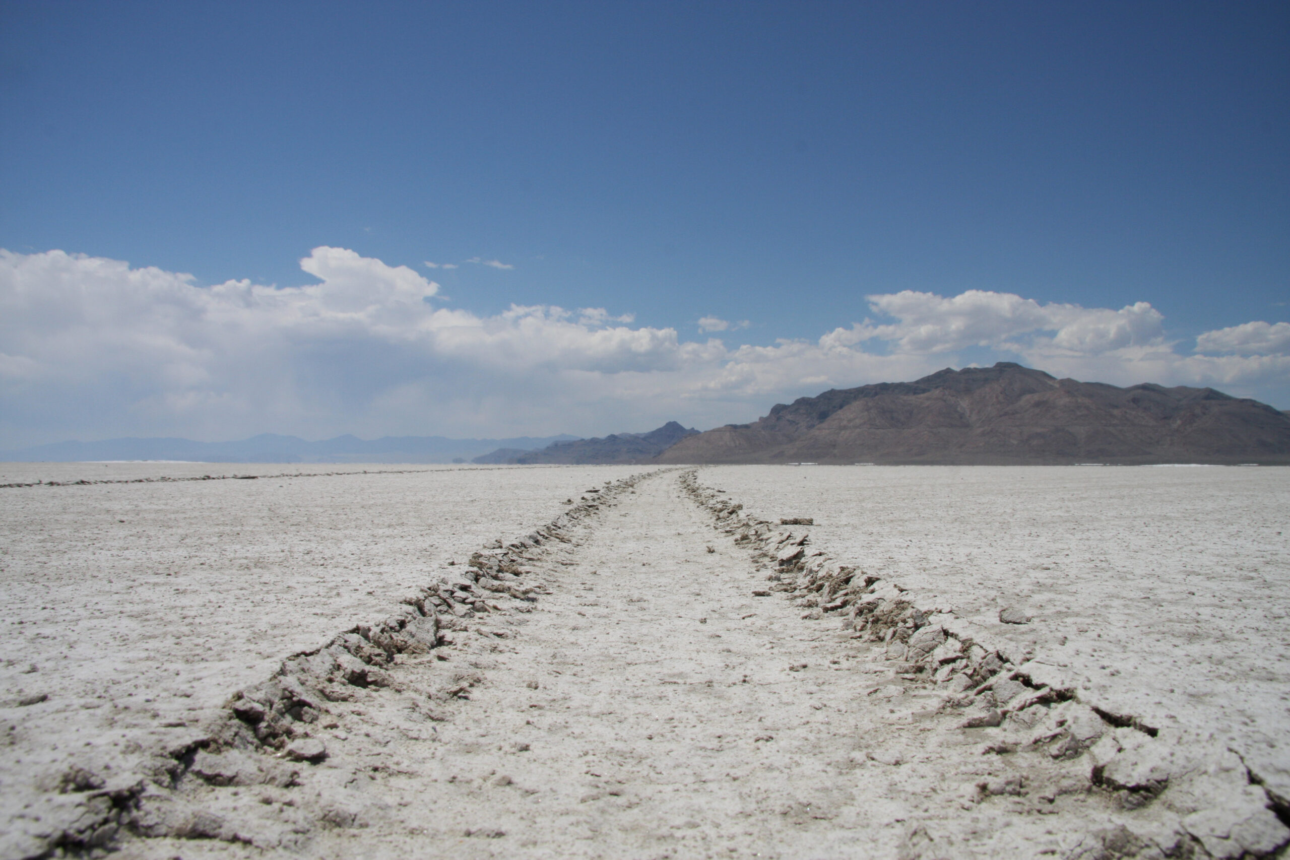 there is a road which goes off into the distance eventually meeting a mountain range. The ground is covered in snow, and the sky is mostly clear with some clouds in the distance.