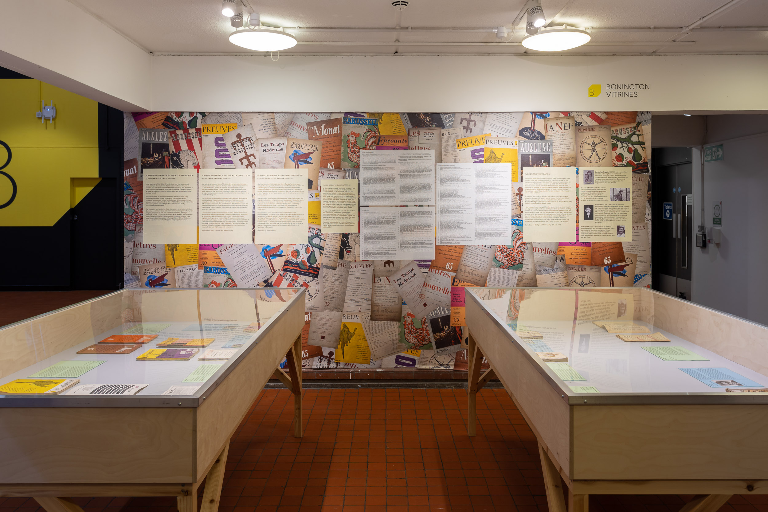 installation view of an exhiition, two large display cases in the foreground, the wall is covered in colourful print outs of magazine covers with text displays on top