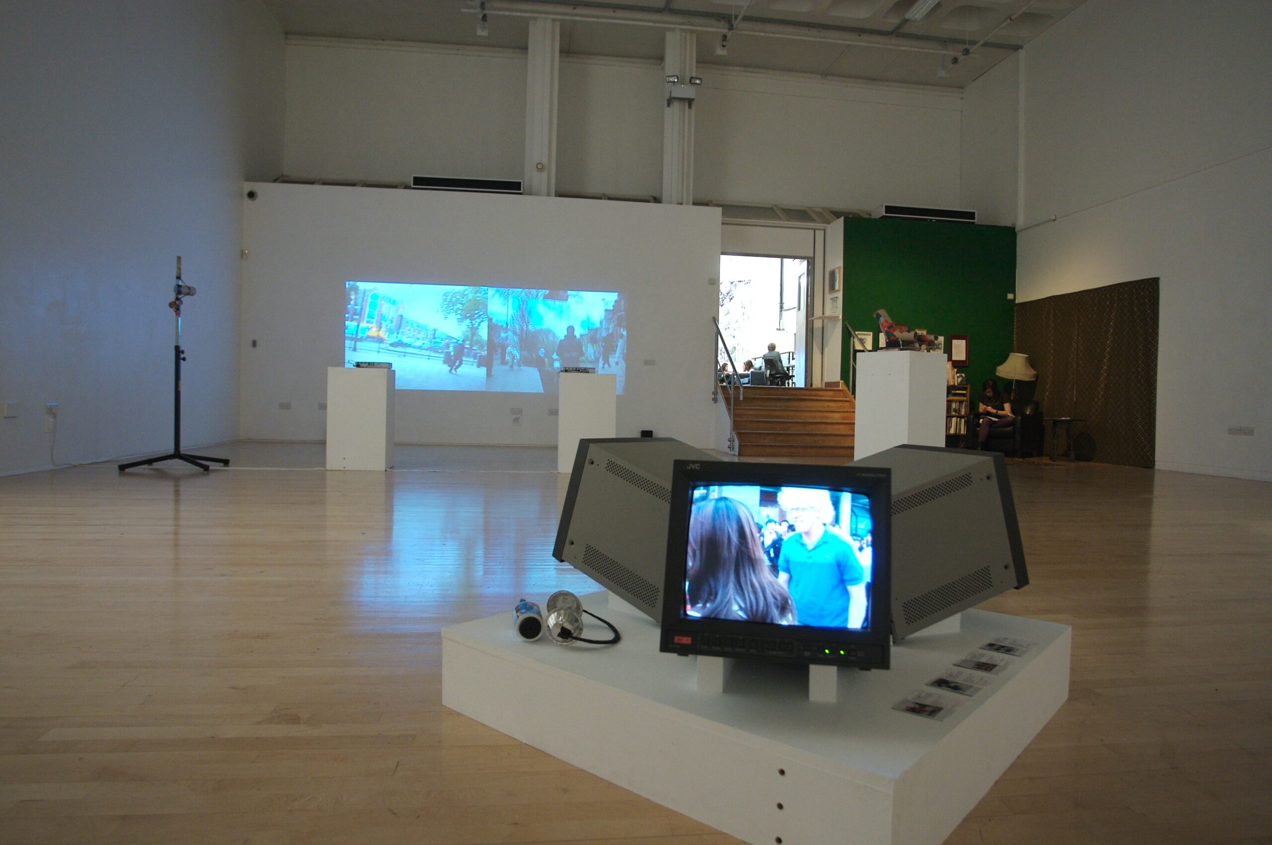 Shows an exhibition view, with a close up of an old CRT tv on a plinth with two other screens. In the back ground there is a projection on the wall.