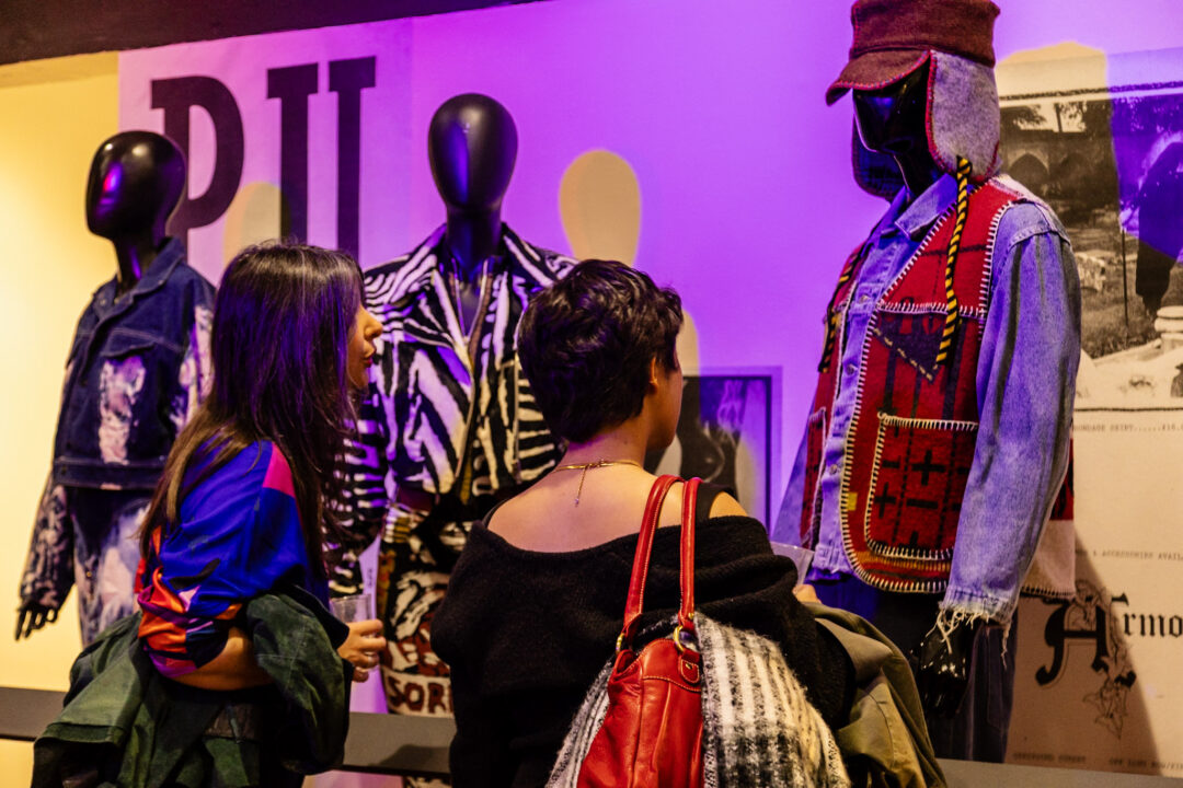 Two people looking at an exhibition of clothing, featuring 3 mannequins wearing 80s clothing.
