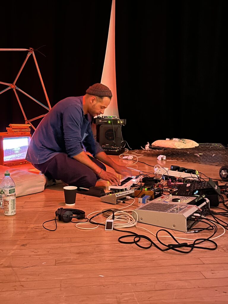 Tom Harris doing a live electronic music performance. He is knelt on the floor, surrounded by different types of sound equipment