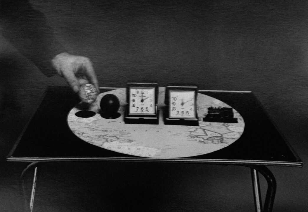 A table with a world map design, featuring two clocks both showing 7:55, a small globe, a black spherical object, and a small model. A hand is reaching toward the globe.
