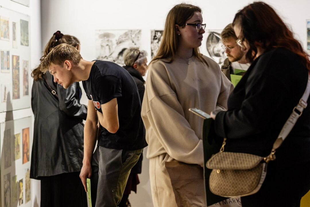 Visitors in an art gallery viewing and discussing various artworks displayed on the walls, with some holding exhibition guides or catalogues.
