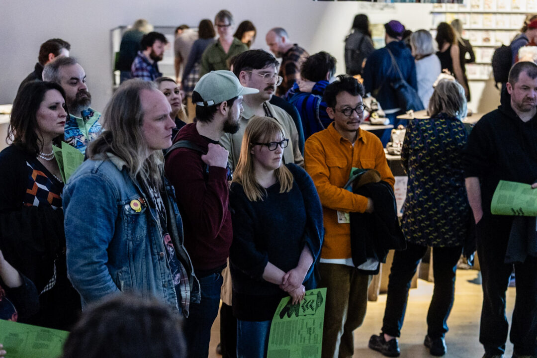 People gathered indoors at bonington gallery, holding green programs, with display tables and artworks in the background.