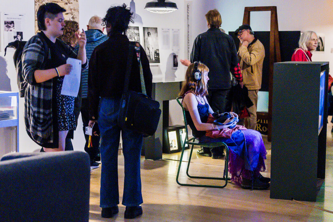 Person wearing headphones sits on a chair at an art exhibit while others stand and view photographs and displays around the room.