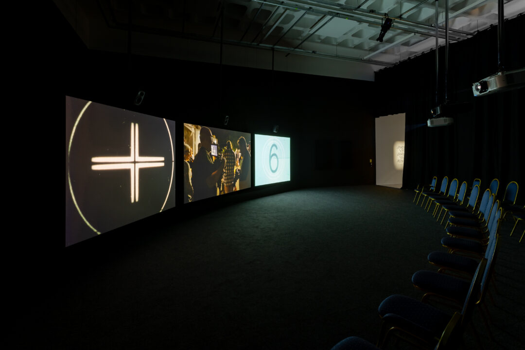 An installation view from inside Bonington Gallery, showing 3 large projection screens positioning in front of an ark of seating. The film is THROUGH A MIRROR, DARKLY by Naeem Mohaiemen.
