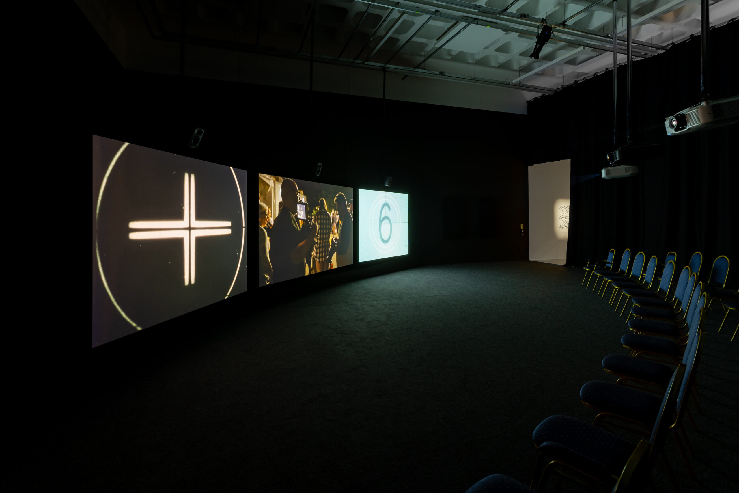 An installation view from inside Bonington Gallery, showing 3 large projection screens positioning in front of an ark of seating. The film is THROUGH A MIRROR, DARKLY by Naeem Mohaiemen.