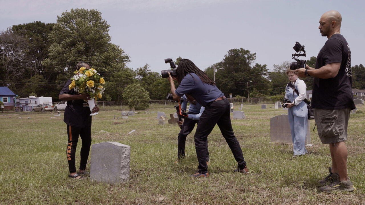 A small group of people stand in an open cemetery with scattered headstones and tall green grass. One individual holds a large bouquet of yellow flowers in front of a headstone. Another person stands close, photographing the scene with a camera. Additional individuals nearby hold video or photography equipment, capturing the moment from different angles. Trees and a few small buildings are visible in the background under a bright sky.