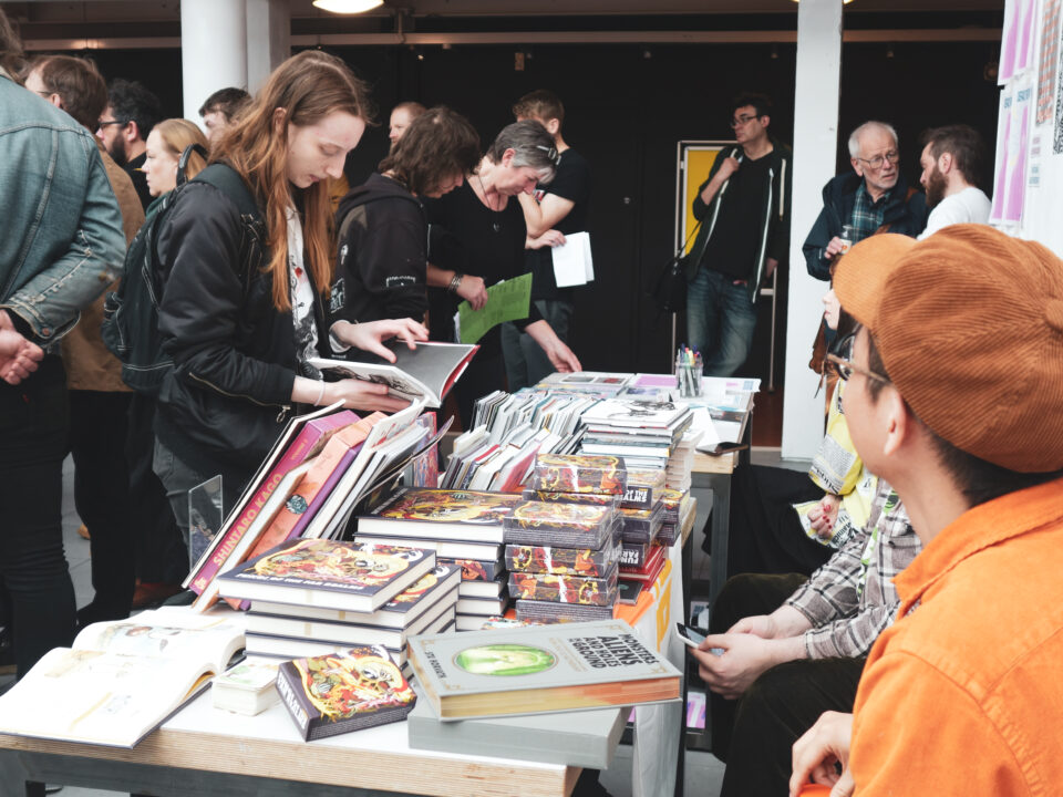 A busy indoor book fair or market scene with several people gathered around tables covered in stacks of books and printed materials. In the foreground, an open book lies on a table surrounded by piles of colourful paperbacks and hardcovers. A person in an orange cap is seated behind the table, appearing to speak with attendees. More people stand closely together in the background, browsing, reading, and discussing books. The setting has a casual, lively atmosphere with posters and displays visible on the walls.